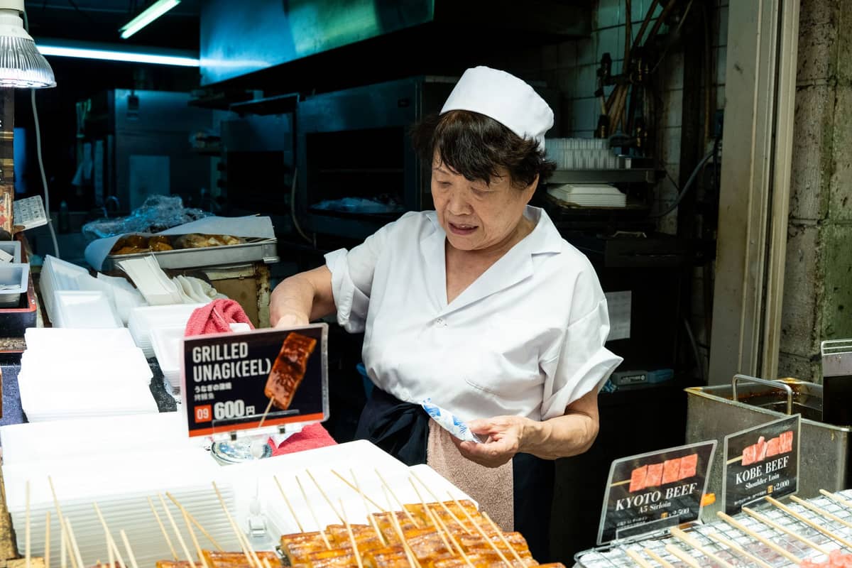 Woman preparing and selling skewered street food including grilled unagi and Kyoto beef in a market food stall
