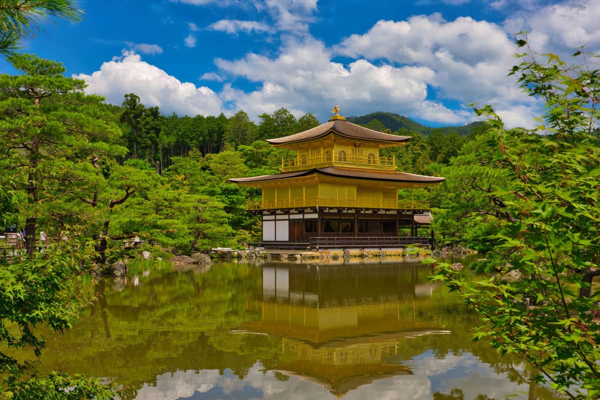 Kinkaku-ji, the iconic Golden Pavilion, reflected in a serene pond surrounded by lush greenery and trees under a bright blue sky in Kyoto.