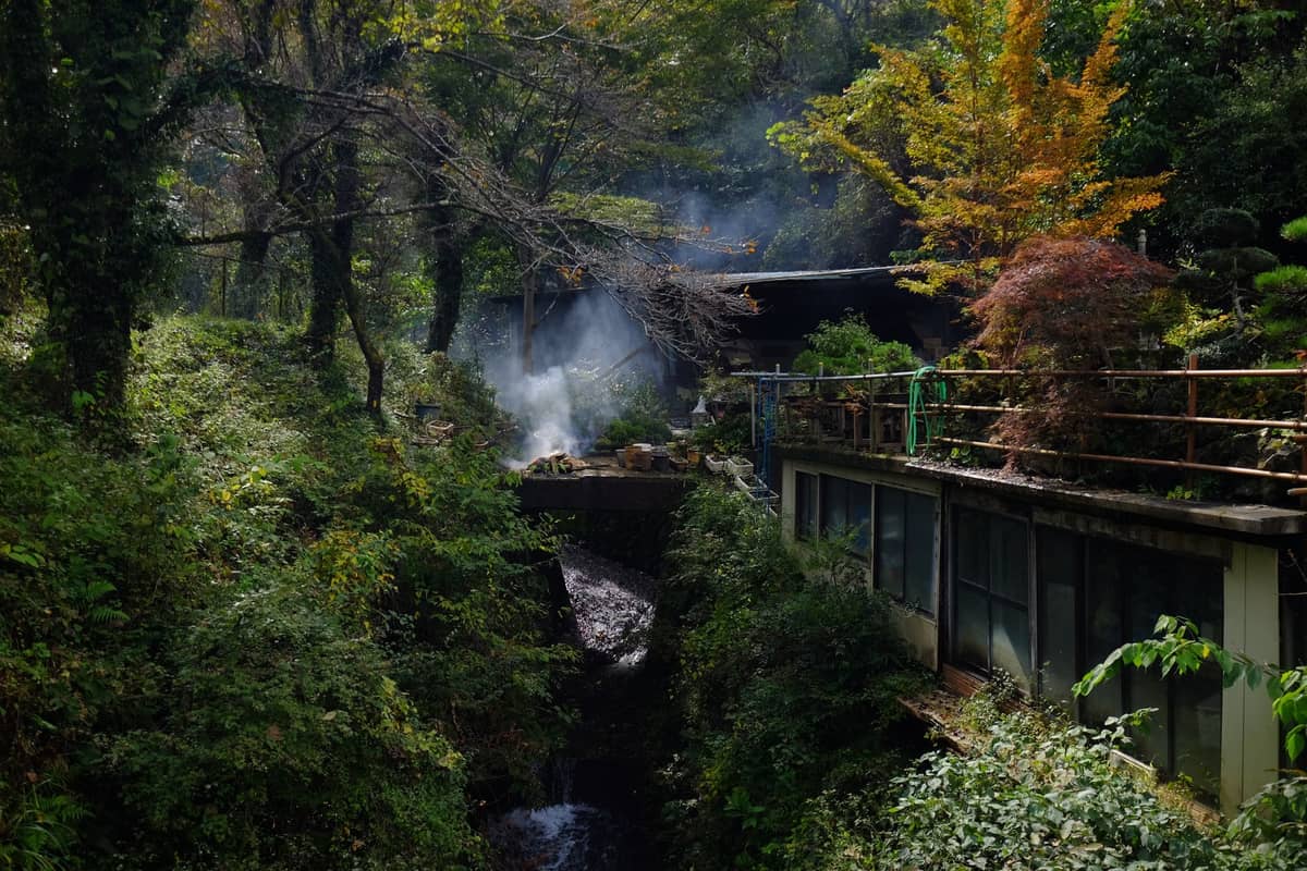 Forest setting with an onsen building, rising steam, and autumn foliage beside a narrow ravine in the Kurama mountains.