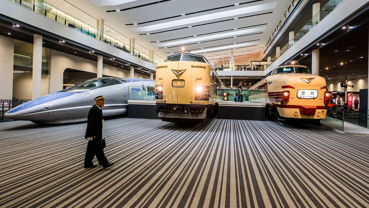 The main train hall inside the Kyoto Railway Museum displaying an original Shinkansen, vintage yellow and cream trains, and a staff member walking past the exhibits.