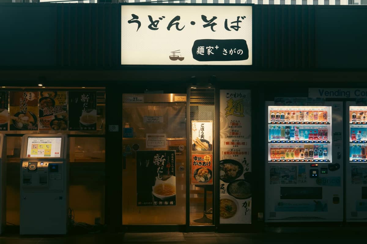 Exterior of a Japanese noodle restaurant at night with vending machines and a ticket machine for ordering food visible outside.