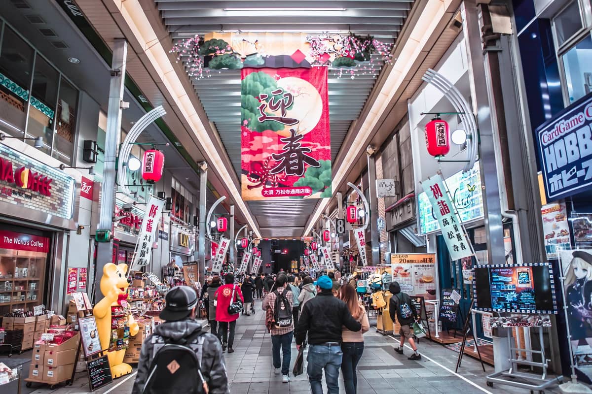 Busy indoor Kyoto shopping arcade with people walking, colorful banners, shops, and a festive market atmosphere.