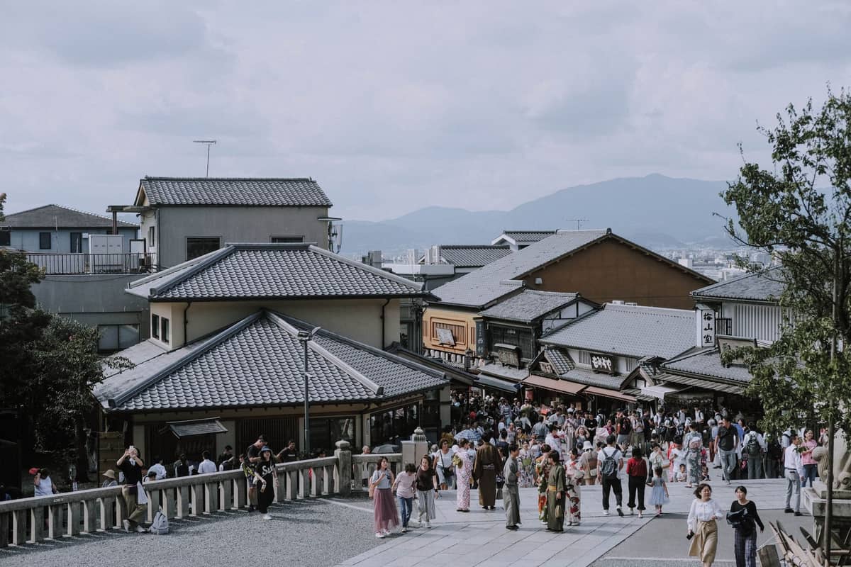 Busy traditional street in Kyoto lined with shops and crowds, some people wearing kimono, with tiled rooftops and distant mountains visible.