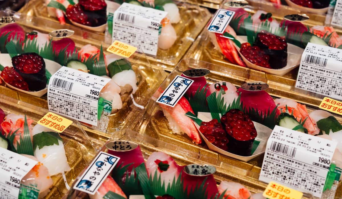 Multiple packs of assorted sushi in plastic trays displayed for sale at a Japanese market with price tags and labels.