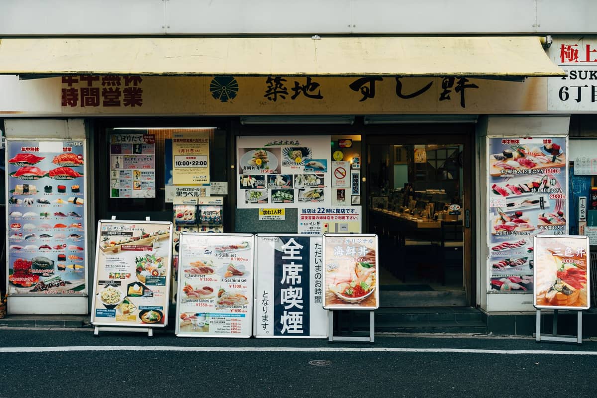 Street view of a sushi restaurant in Japan with colorful menu boards and food displays outside the entrance.
