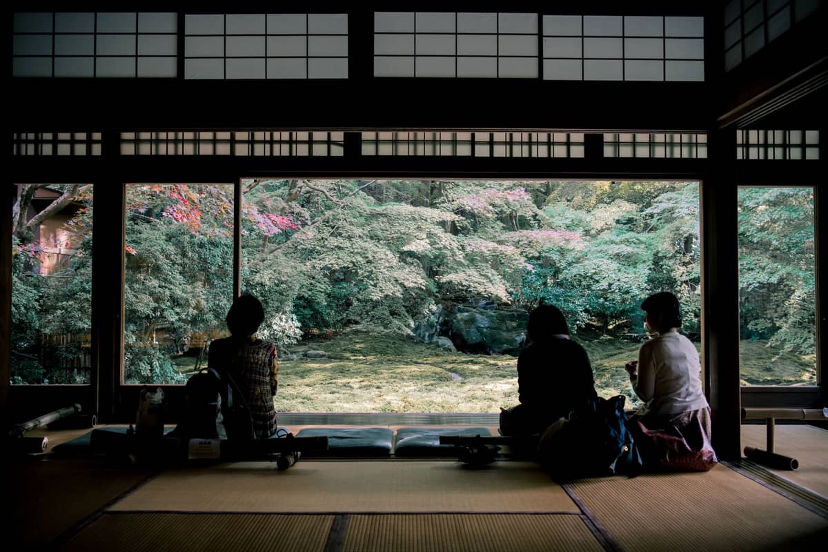 Three people sit on tatami mats inside a traditional Japanese room, gazing out at a lush garden through large open windows.