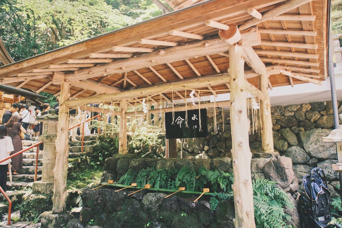 Chozuya pavilion at a Shinto shrine with ladles for purification, some visitors nearby, and lush greenery in the background.