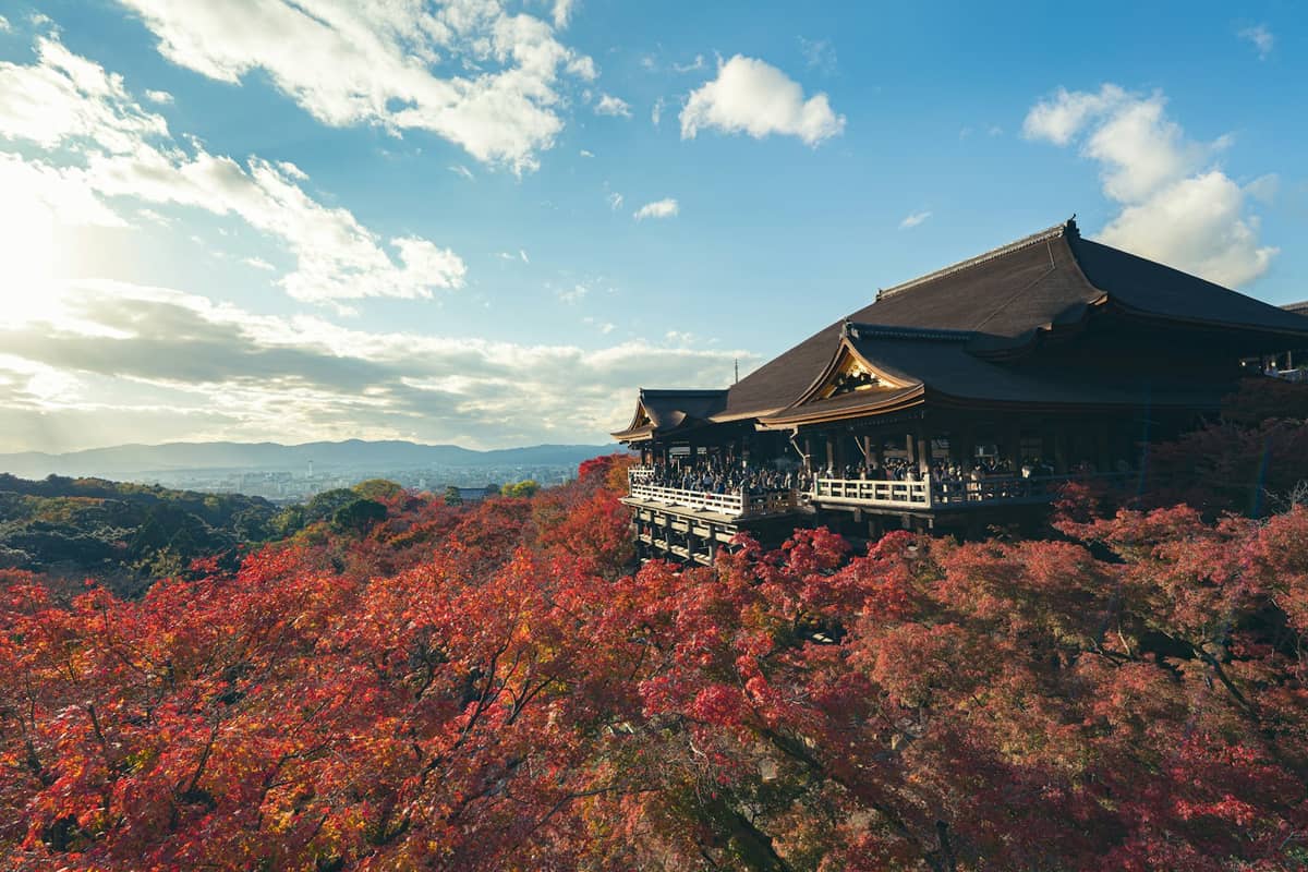 Kiyomizu-dera temple in Kyoto framed by vivid autumn foliage under a blue sky, with crowds visible on the terrace.