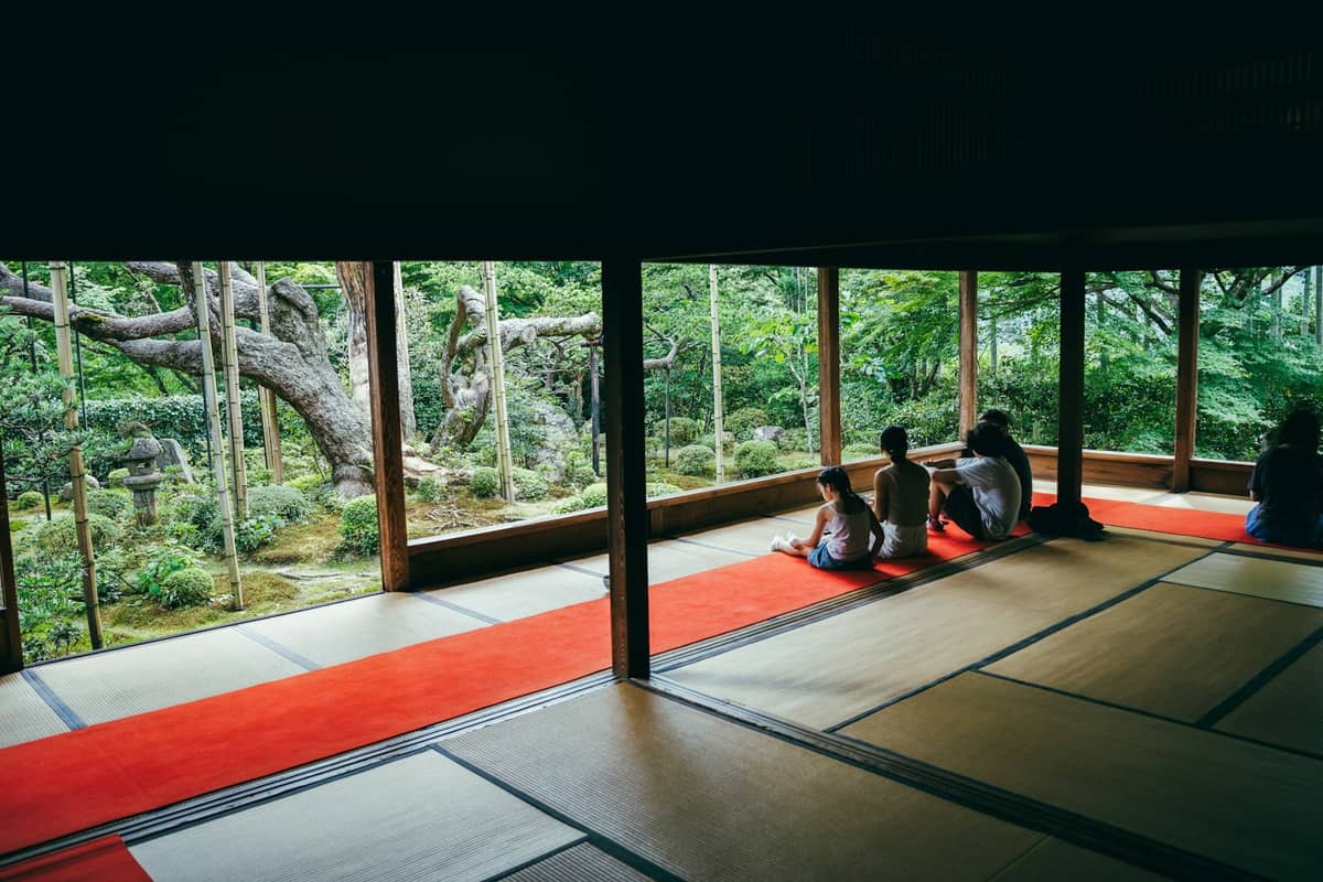 Tatami room in a Kyoto Buddhist temple with people quietly sitting by large windows, overlooking a tranquil green garden.