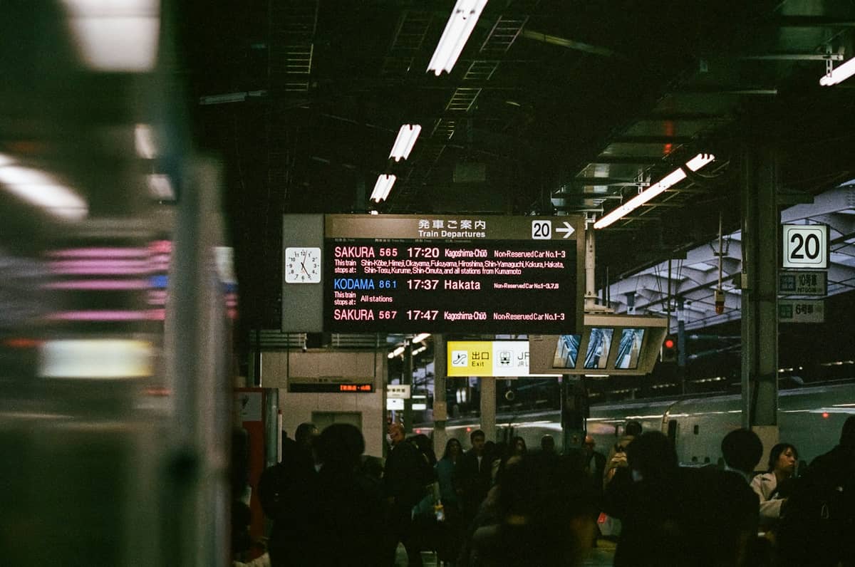 Crowded platform at a Japanese train station with digital signs above, displaying Shinkansen departure information in Japanese and English.