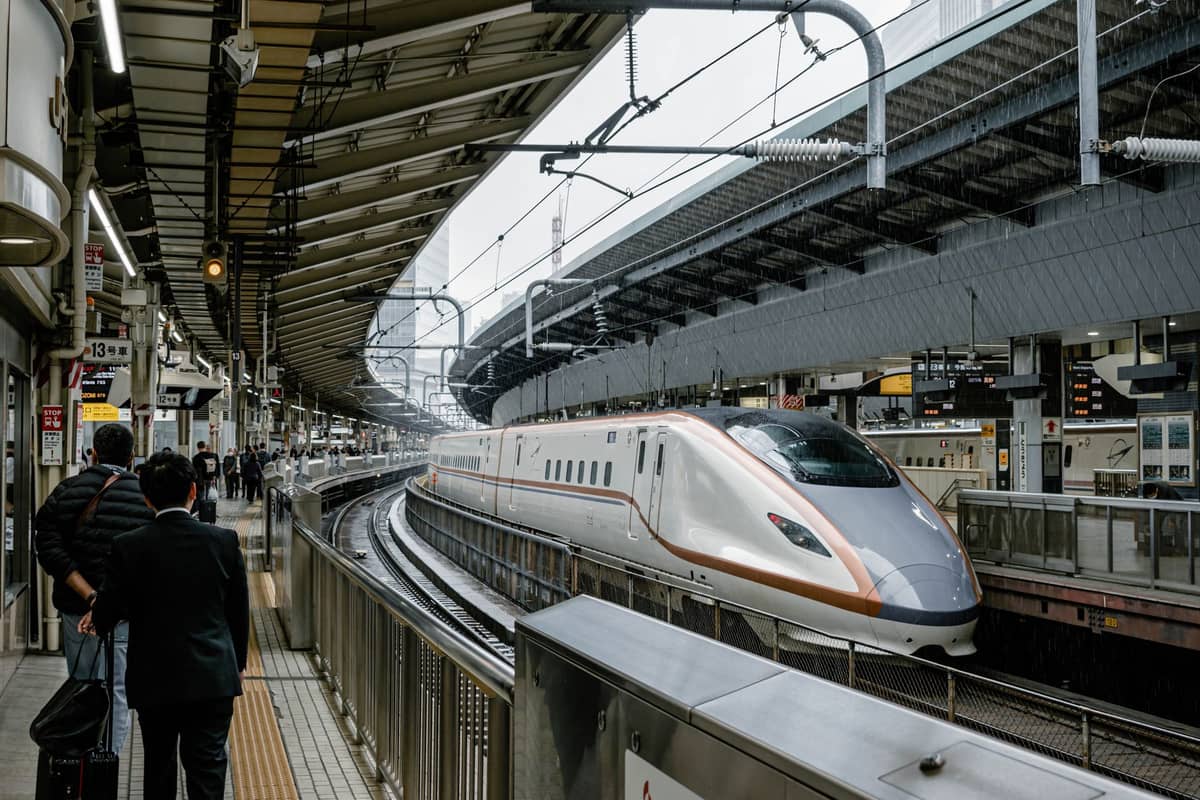 A modern shinkansen bullet train at a platform in a major Japanese station, with passengers waiting and overhead signs visible in the background.