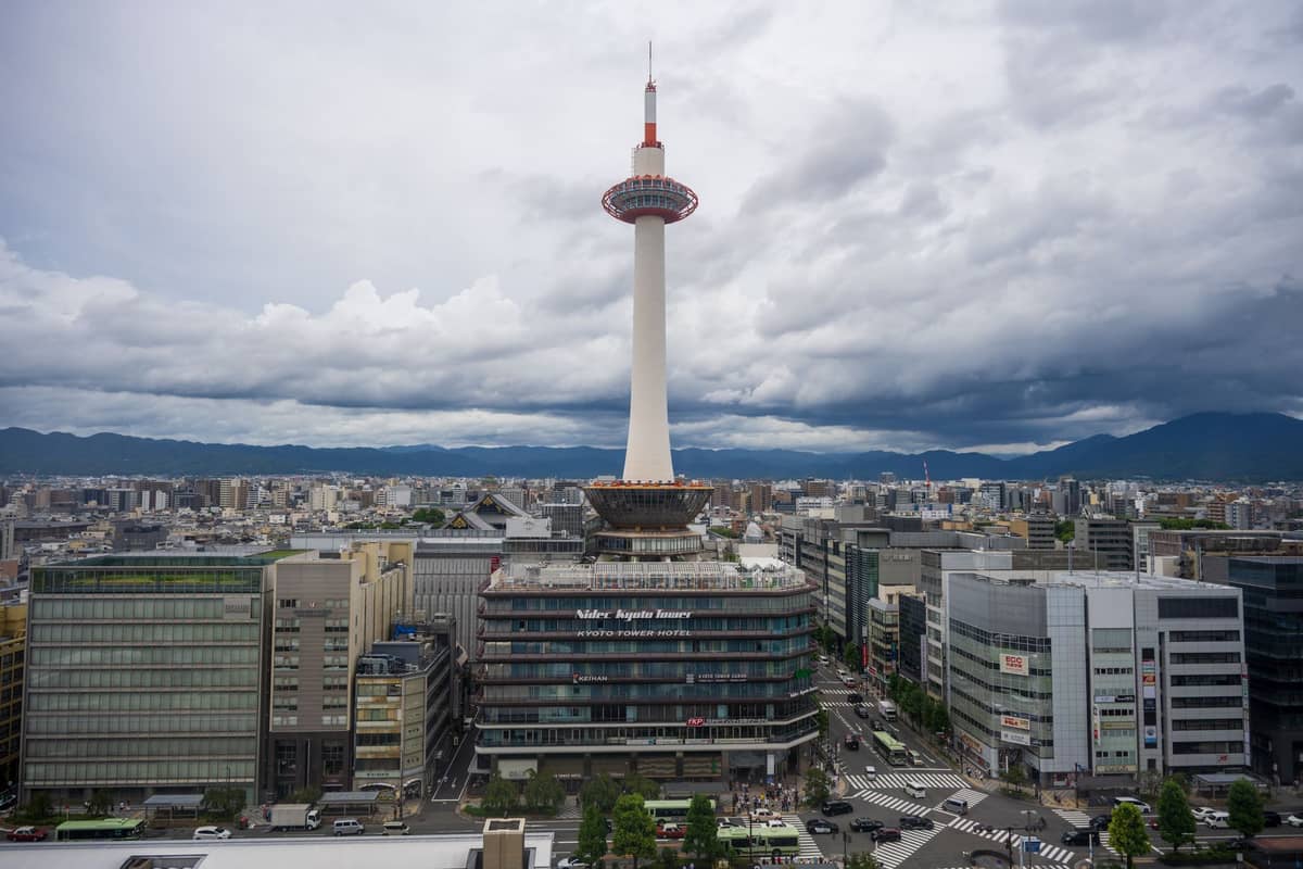 Kyoto Tower rising above neighboring buildings in downtown Kyoto, with a dramatic cloudy sky and mountains in the background.