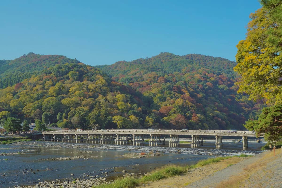 A wide view of the Togetsukyo Bridge crossing the Katsura River with lush, forested mountains in Arashiyama, Kyoto, under a clear blue sky.