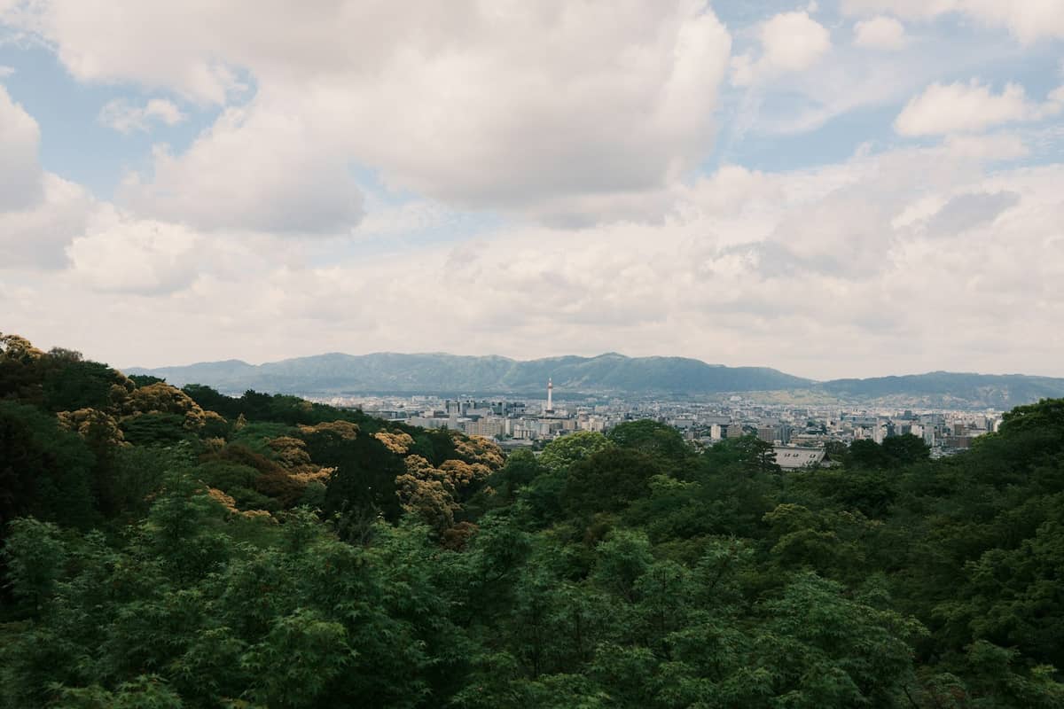 Landscape view of Kyoto city with mountains surrounding the basin, seen under a partly cloudy sky and greenery in the foreground.