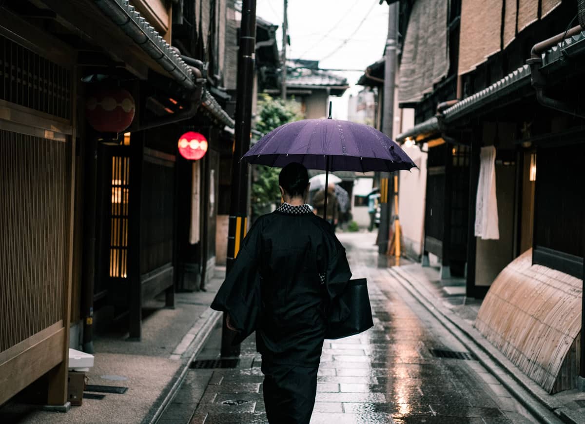 Person in traditional clothing walks with an umbrella down a wet, narrow street lined with Kyoto-style wooden buildings after rainfall.
