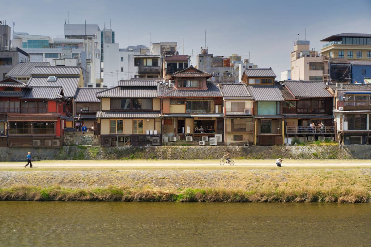 Riverside pathway in Kyoto with traditional wooden houses, a cyclist, and a pedestrian, illustrating outdoor travel options in good weather.