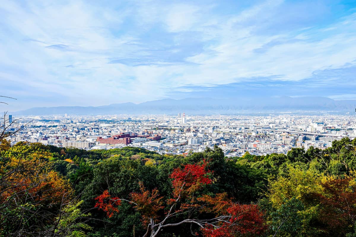 Wide landscape view of Kyoto city with autumn trees and bright sky, showing the cityscape surrounded by mountains and colorful seasonal foliage.