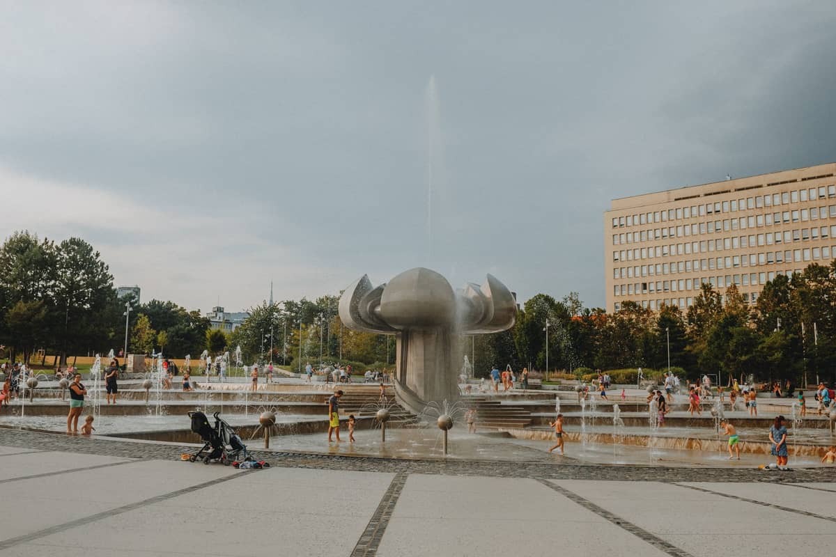 Children playing in a large modern fountain and splash area with a stroller and parents nearby, city building and greenery in background.