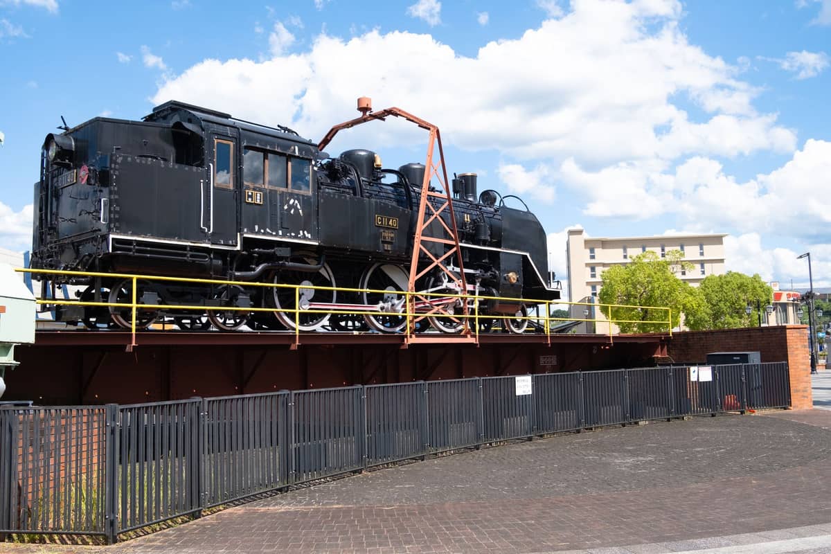 Outdoor display of a black steam locomotive on a turntable at Kyoto Railway Museum, with blue sky and city buildings in the background.