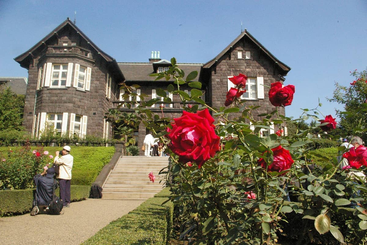 A grand Western-style brick mansion with white windows stands behind vibrant red roses in the Kyu-Furukawa Gardens under a clear blue sky.