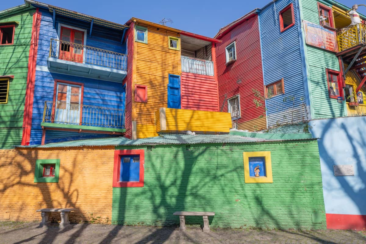บ้านสังกะสีทาสีสด ๆ เรียงรายริมถนนใน Caminito, La Boca บัวโนสไอเรส มีม้านั่งและเงาต้นไม้อยู่เบื้องหน้า