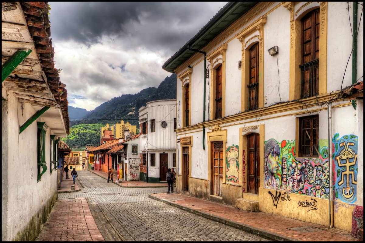 Colorful colonial street scene in Bogotá’s La Candelaria, showing historic buildings, murals, and the green foothills of the Andes in the background.