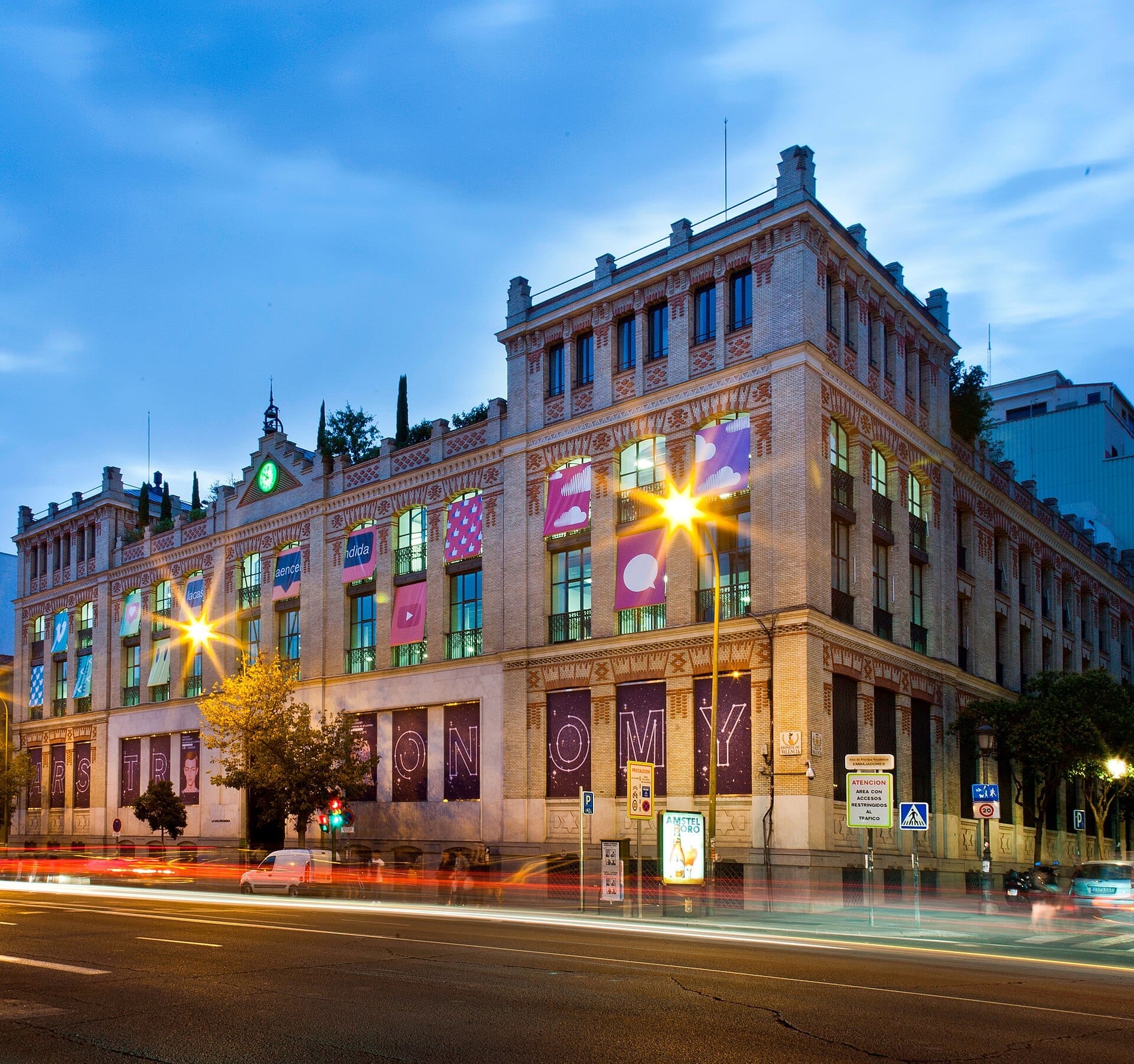 La facciata neo-mudéjar di La Casa Encendida a Madrid illuminata al crepuscolo, con striscioni colorati e traffico cittadino sfocato in primo piano.