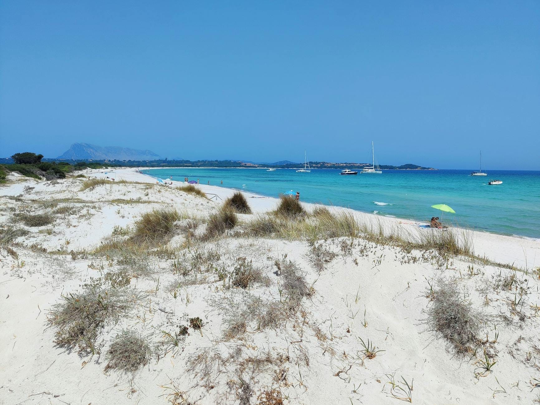 Weitblick über La Cinta Beach mit feinen weißen Sanddünen im Vordergrund und türkisfarbenem Wasser, in dem Segelboote unter einem klaren blauen Himmel vor Anker liegen.