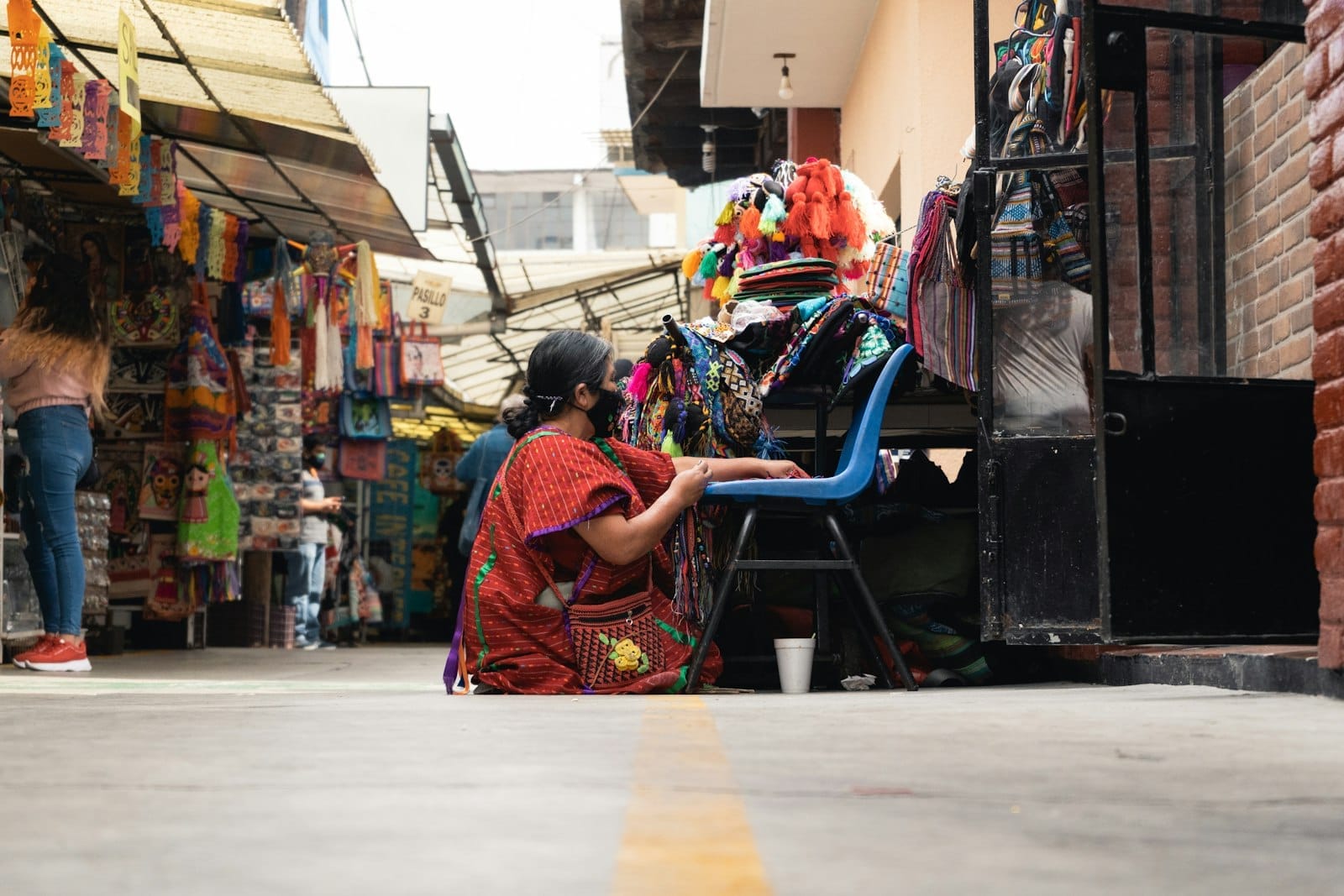 Una donna in abiti tradizionali siede in un vivace mercato artigianale, circondata da colorate creazioni fatte a mano e tessuti esposti lungo le bancarelle.