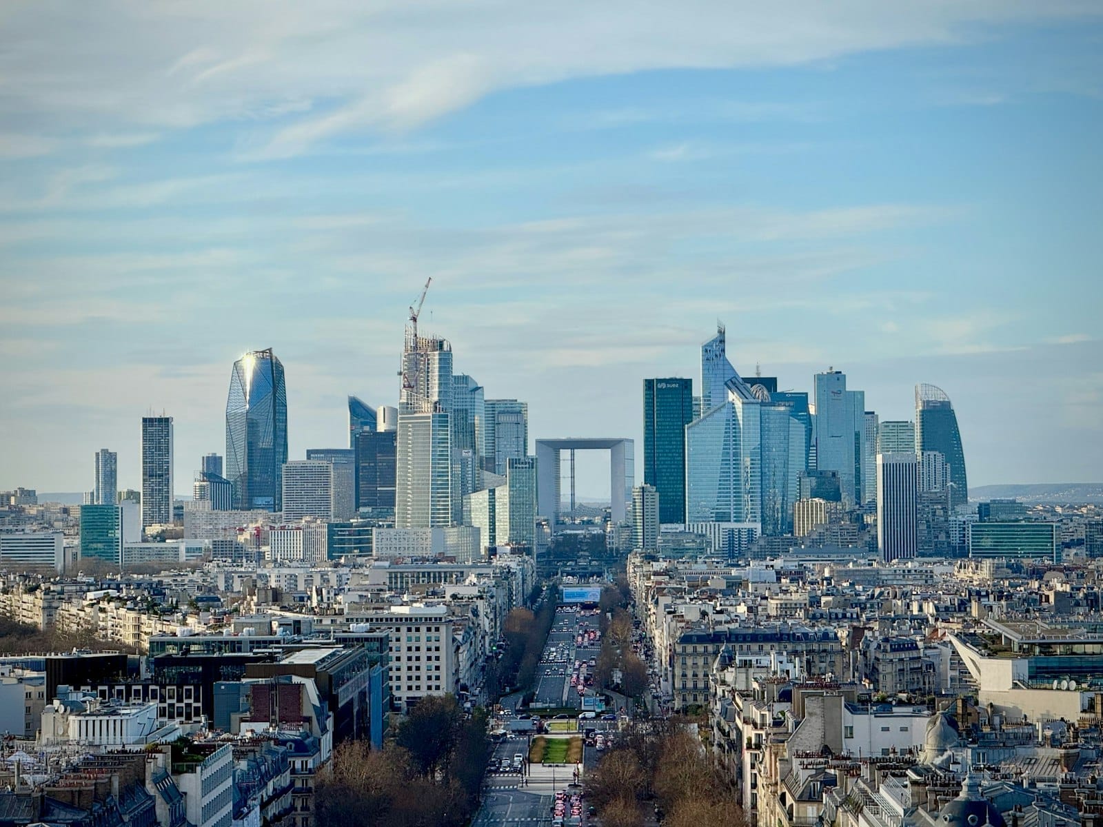 Un'ampia veduta di La Défense con i grattacieli di vetro e la Grande Arche, vista in lontananza sopra i tetti di Parigi sotto un cielo azzurro.