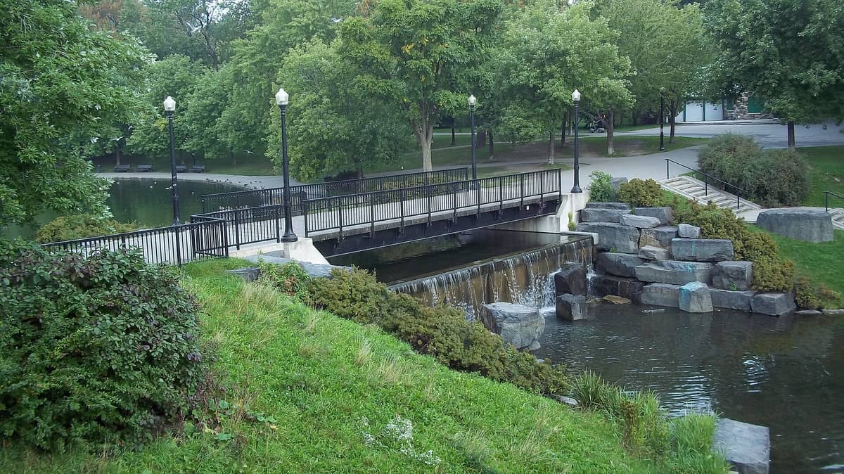 Brücke über einen Teich mit kleinem Wasserfall, umgeben von Bäumen und Grün im Parc La Fontaine, mit Wegen und Parkbänken im Hintergrund.