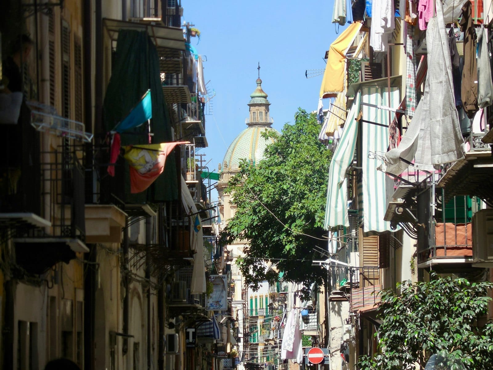 Enge Gasse in La Kalsa, Palermo, gesäumt von alten Gebäuden, bunter Wäsche an den Balkonen und einer Kirche mit Kuppel im Hintergrund.