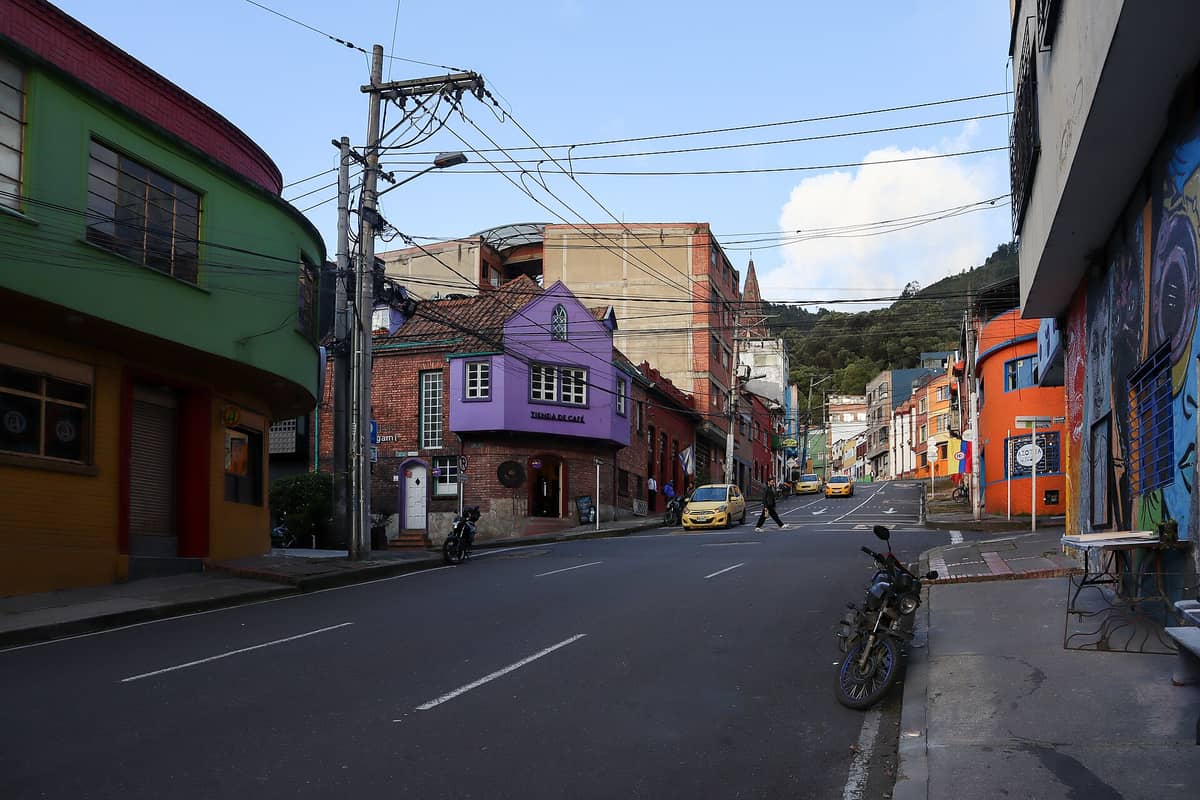 Colorful and eclectic streetscape of La Macarena, Bogotá, with vibrant buildings, murals, cafes, and taxis under a clear sky.