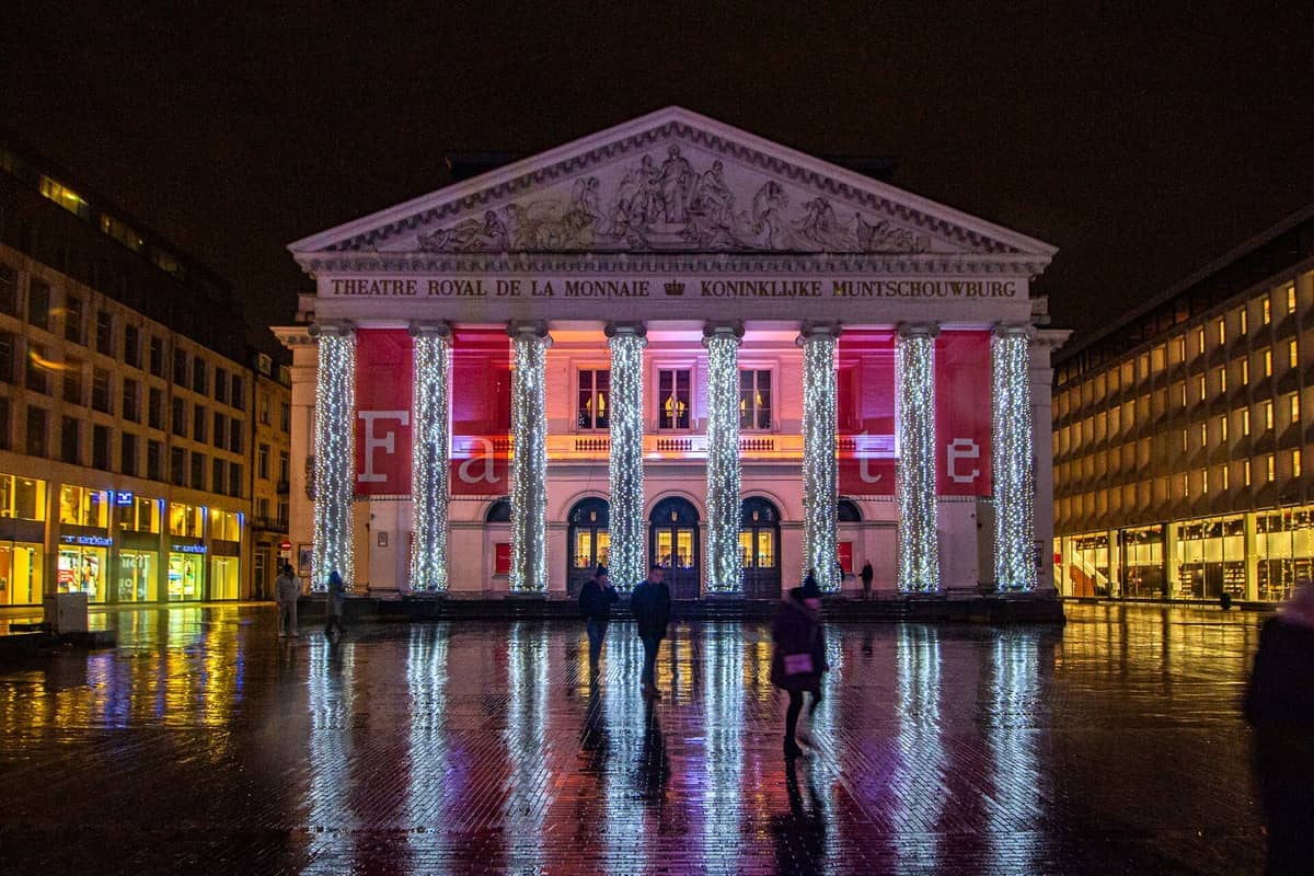 Vista noturna da La Monnaie em Bruxelas com colunas neoclássicas iluminadas e pedestres em uma praça molhada e refletiva.