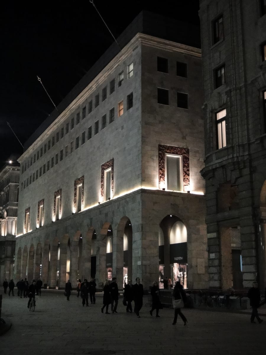 Il grande magazzino La Rinascente Milano illuminato di notte, con facciata in pietra ad archi e clienti serali che camminano lungo Piazza del Duomo.