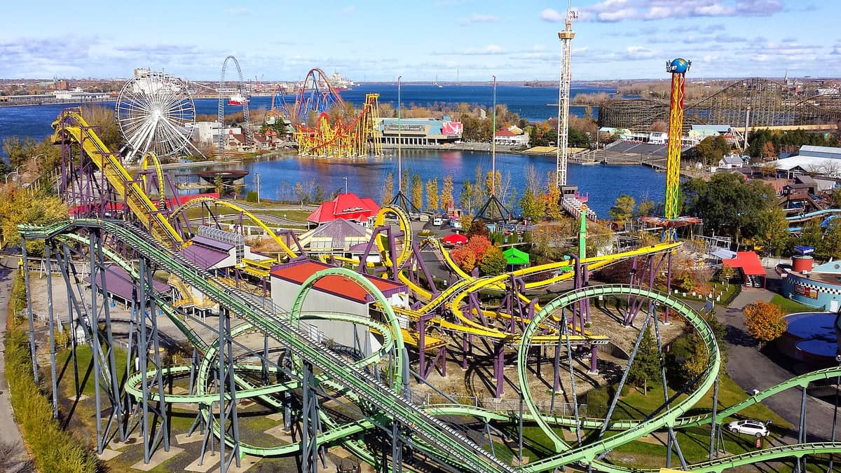 Ein weitläufiger Blick aus der Vogelperspektive auf den Freizeitpark La Ronde in Montreal mit bunten Achterbahnen, einem Riesenrad, Wasser und dem Sankt-Lorenz-Strom im Hintergrund.