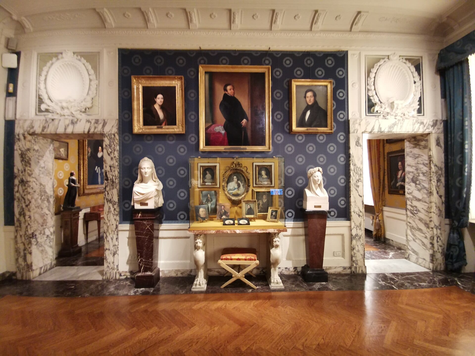 Galleria interna del Museo Teatrale alla Scala con piedistalli in marmo, busti classici, ritratti ornati ed eleganti pareti con motivi blu e luce naturale.