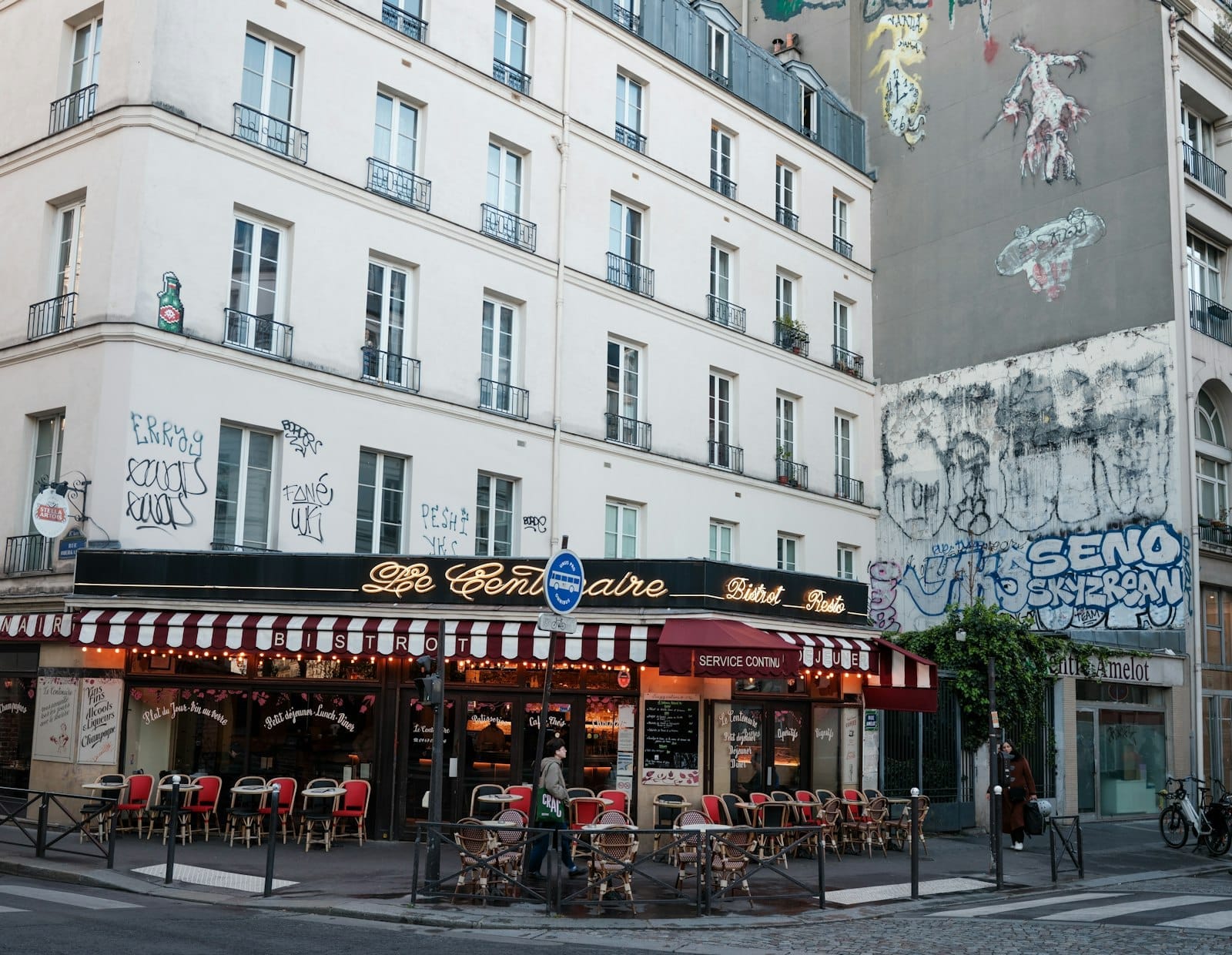 Angolo di strada parigino con un classico café, tavolini all'aperto, street art e graffiti su un edificio vicino, nel vivace 19° arrondissement.