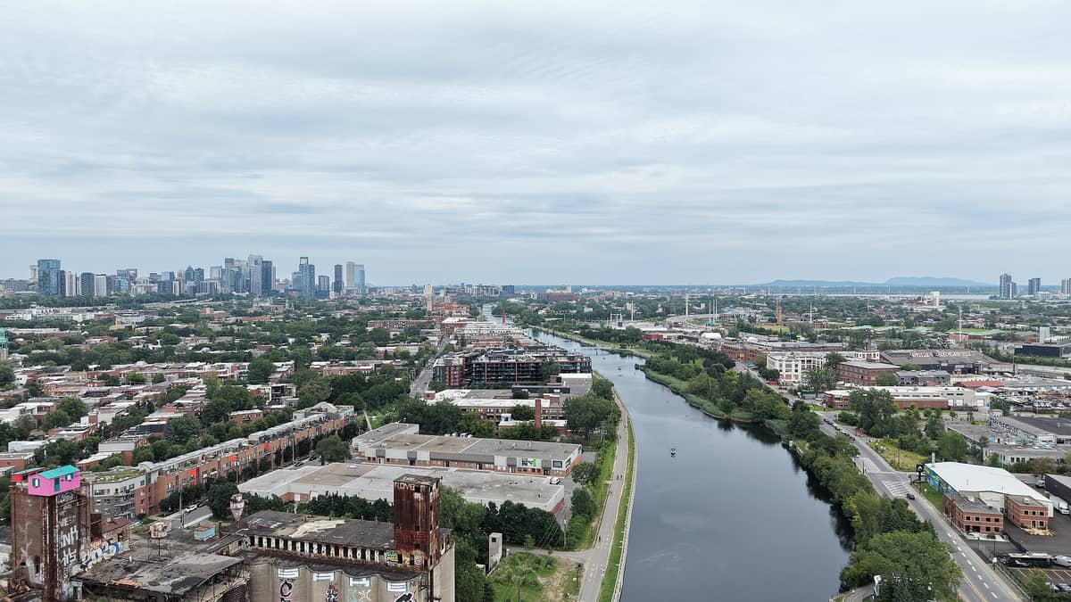 Luftaufnahme des Lachine-Kanals, der durch Montréal führt, mit der Innenstadt-Skyline im Hintergrund, grünen Bäumen, Industriegebäuden und einem Radweg am Ufer.