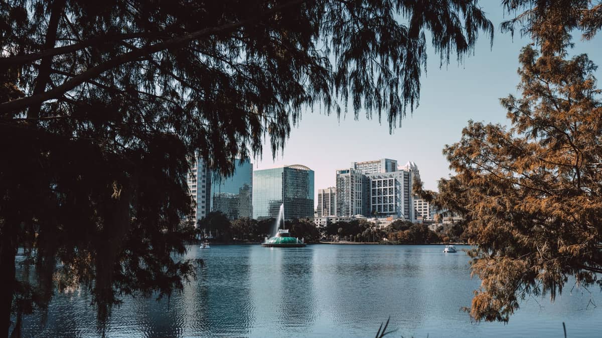 Blick über den Lake Eola mit dem markanten Springbrunnen und der Skyline von Downtown Orlando, eingerahmt von Bäumen im Vordergrund an einem klaren Tag.