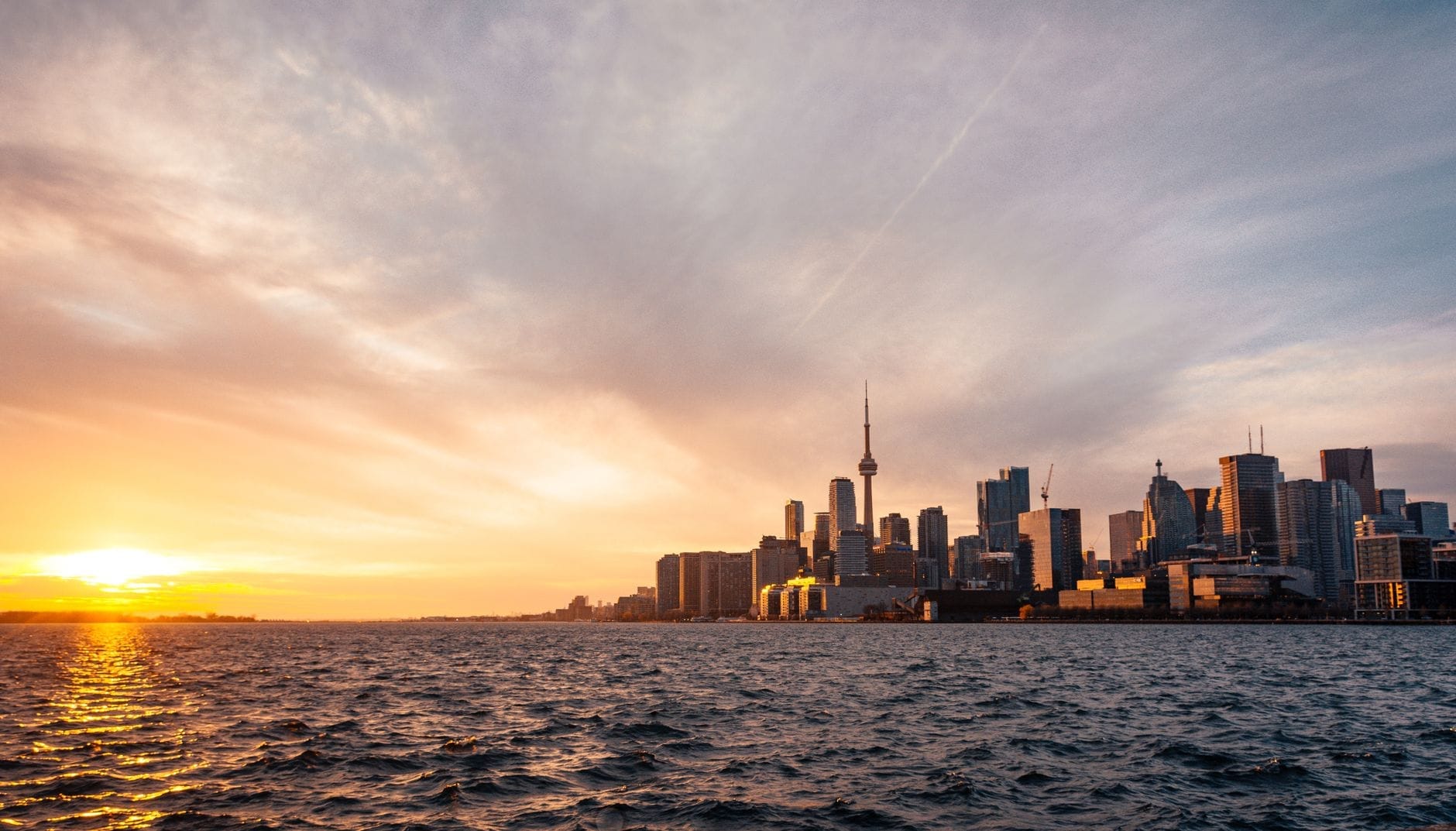 Skyline de Toronto vista do Lago Ontario ao pôr do sol, com os edifícios à beira-mar e a CN Tower em silhueta contra um céu colorido.