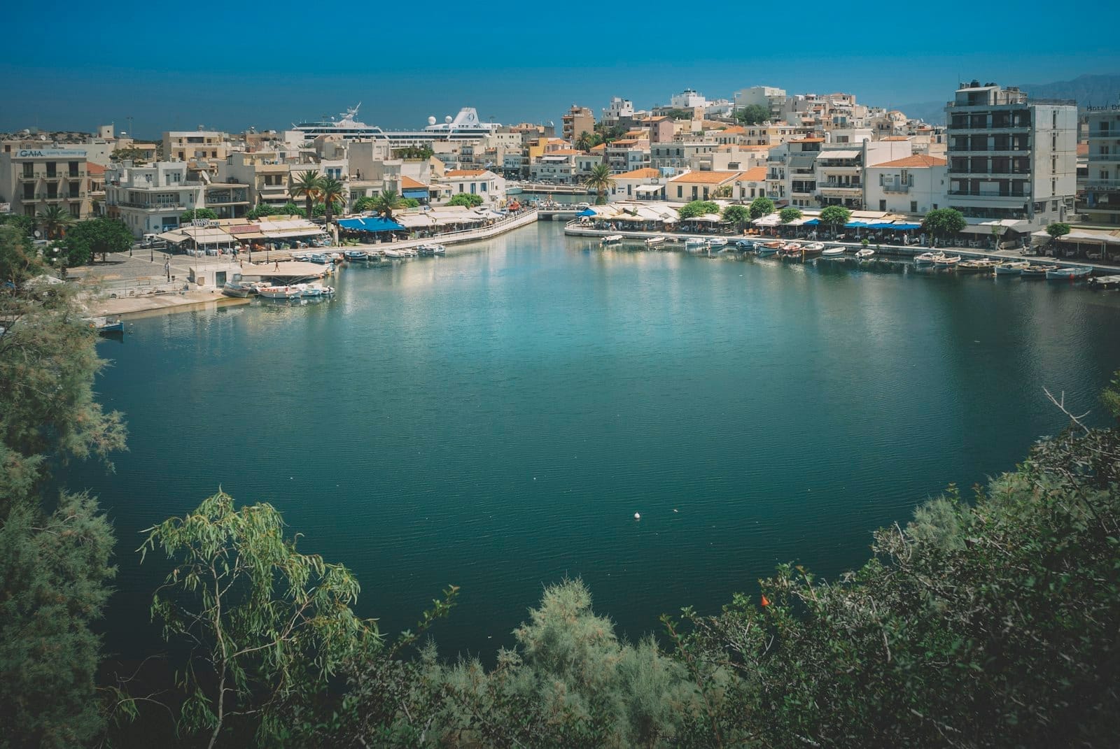 Vista del lago Voulismeni ad Agios Nikolaos con l'acqua blu intenso, i caffè sul lungolago e gli edifici della città che circondano il bacino a forma di cratere sotto un cielo limpido.