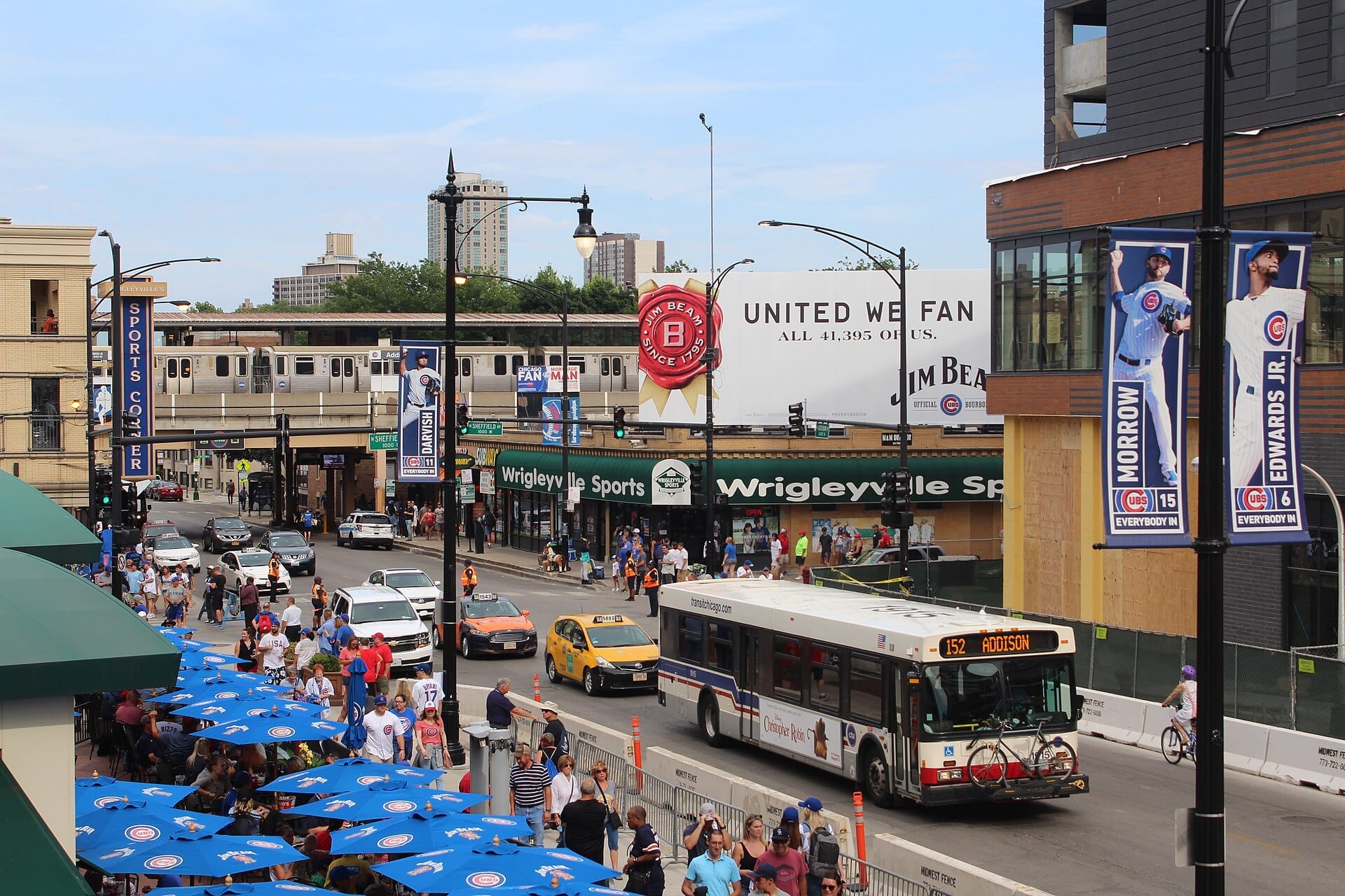 ทางแยกที่คึกคักของ Wrigleyville ใน Lakeview พร้อมฝูงชน รถบัส CTA แท็กซี่ และรถไฟยกระดับข้างๆ ร้านอาหารและบาร์กีฬาในวันที่แดดจ้า