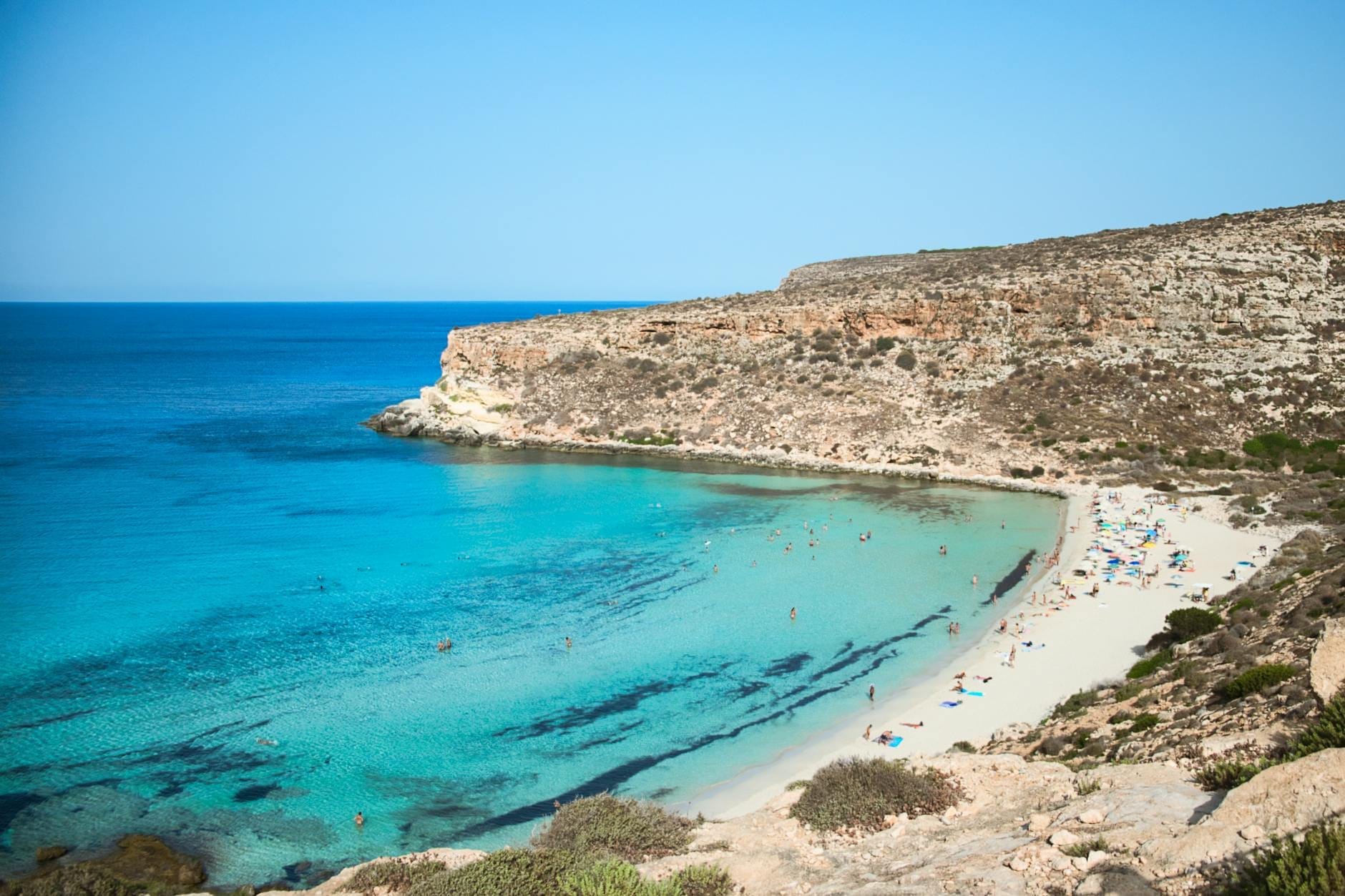 Weitläufiger Blick auf die Spiaggia dei Conigli mit leuchtendem türkisfarbenem Wasser, weißem Sandstrand, sonnenbadenden Besuchern und Felsenklippen unter strahlend blauem Himmel auf Lampedusa.