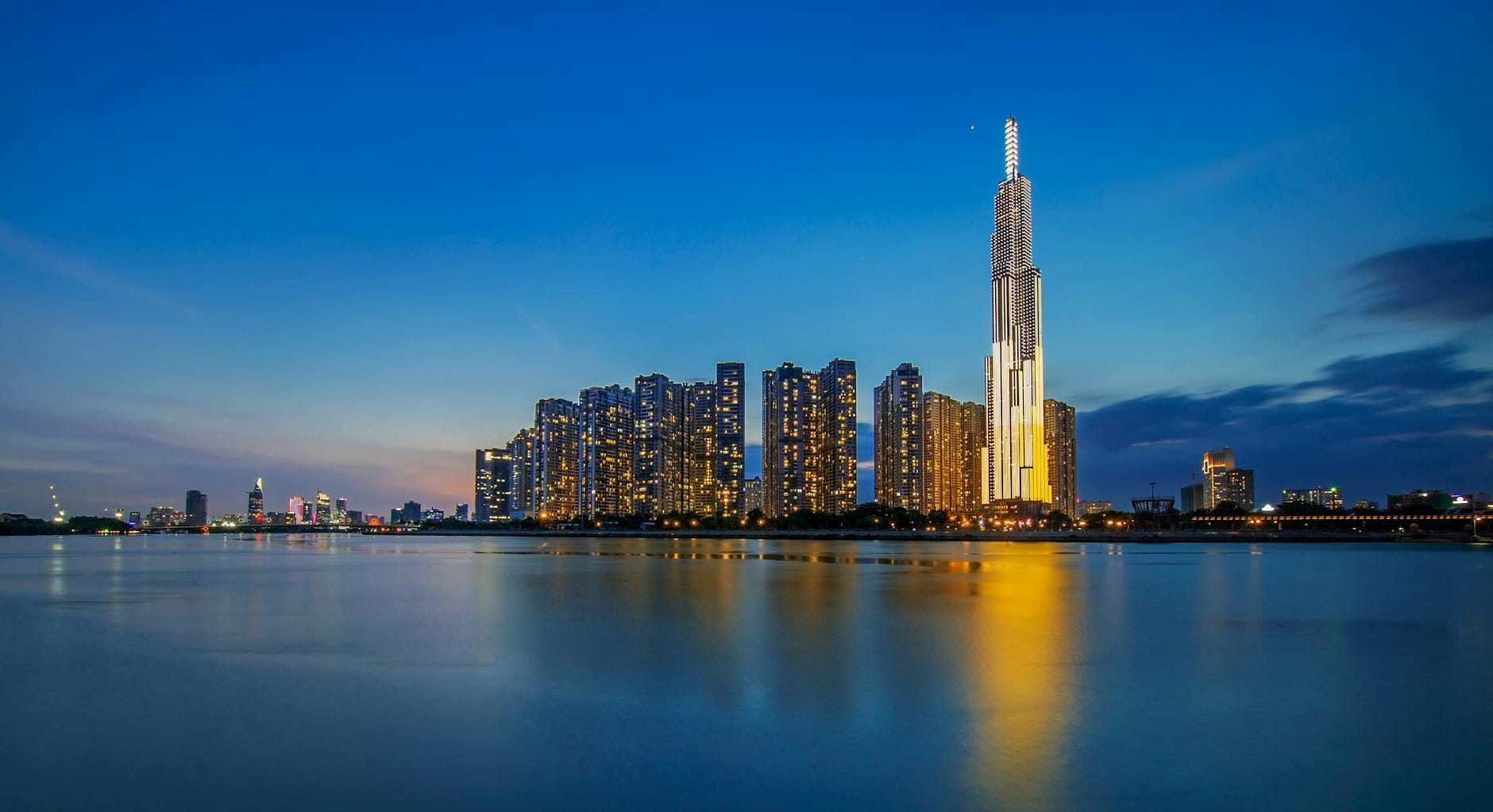 El Landmark 81 iluminado intensamente al atardecer, dominando el skyline y reflejándose en las tranquilas aguas del río bajo un cielo azul vibrante.