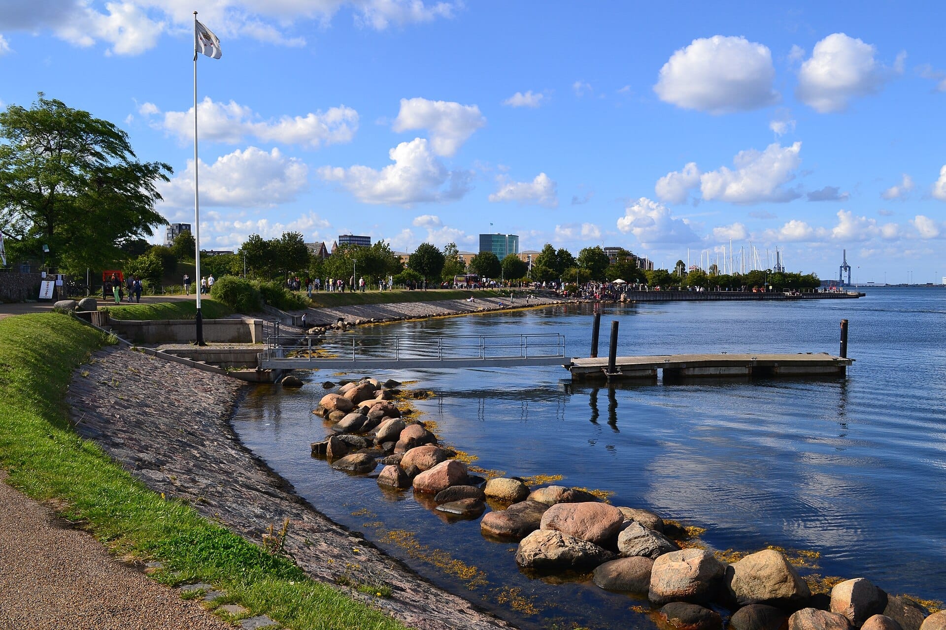Vista della Passeggiata Langelinie a Copenaghen in una giornata di sole, con persone che camminano lungo il lungomare, alberi verdi e acque calme del porto.