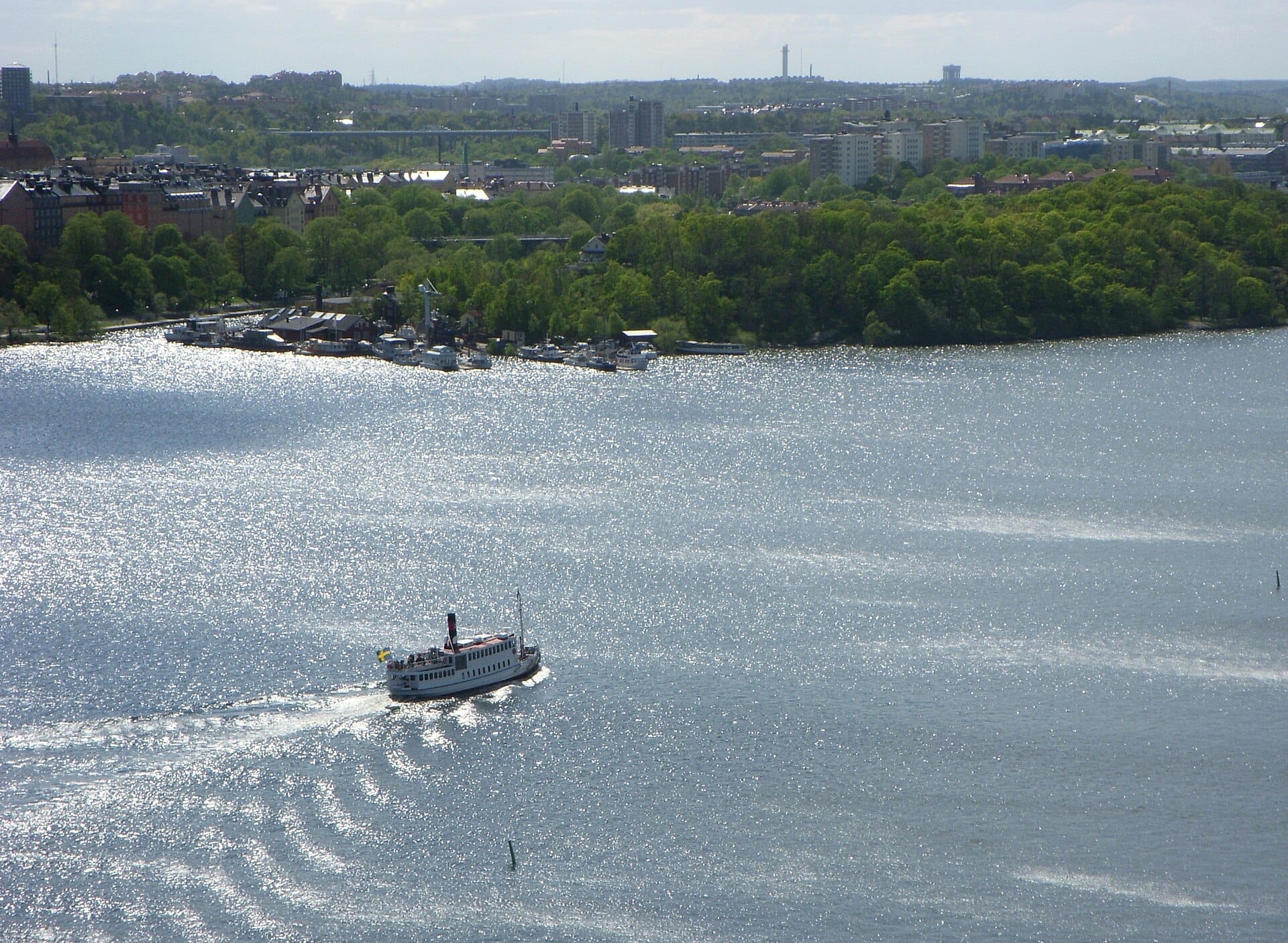 เรือเฟอร์รี่แล่นผ่านชายฝั่งสีเขียวชอุ่มของเกาะ Långholmen โดยมีอาคารในเมือง Stockholm ปรากฏให้เห็นในฉากหลังที่สดใส