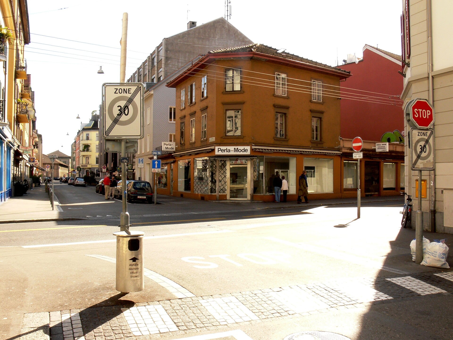 มุมมองถนน Langstrasse ในซูริค มีผู้คนเดินผ่าน ตึกสีน้ำตาลโดดเด่น ร้านค้า ป้ายถนน และแสงแบบเมืองช่วงกลางวัน