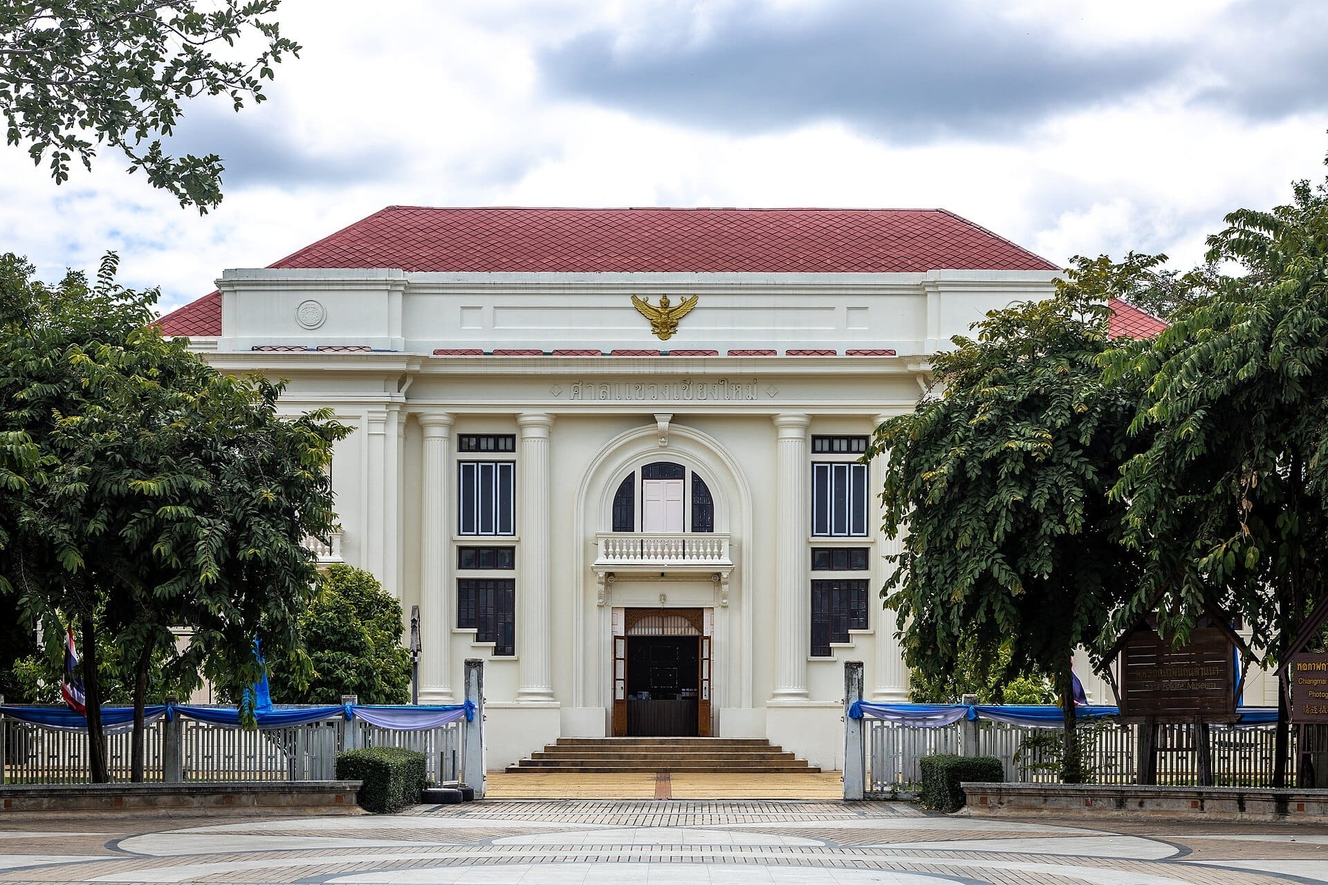 Cảnh nhìn từ phía trước của Lanna Folklife Museum ở Chiang Mai, nổi bật với tòa nhà màu trắng thời thuộc địa, mái đỏ và cây xanh tươi tốt.