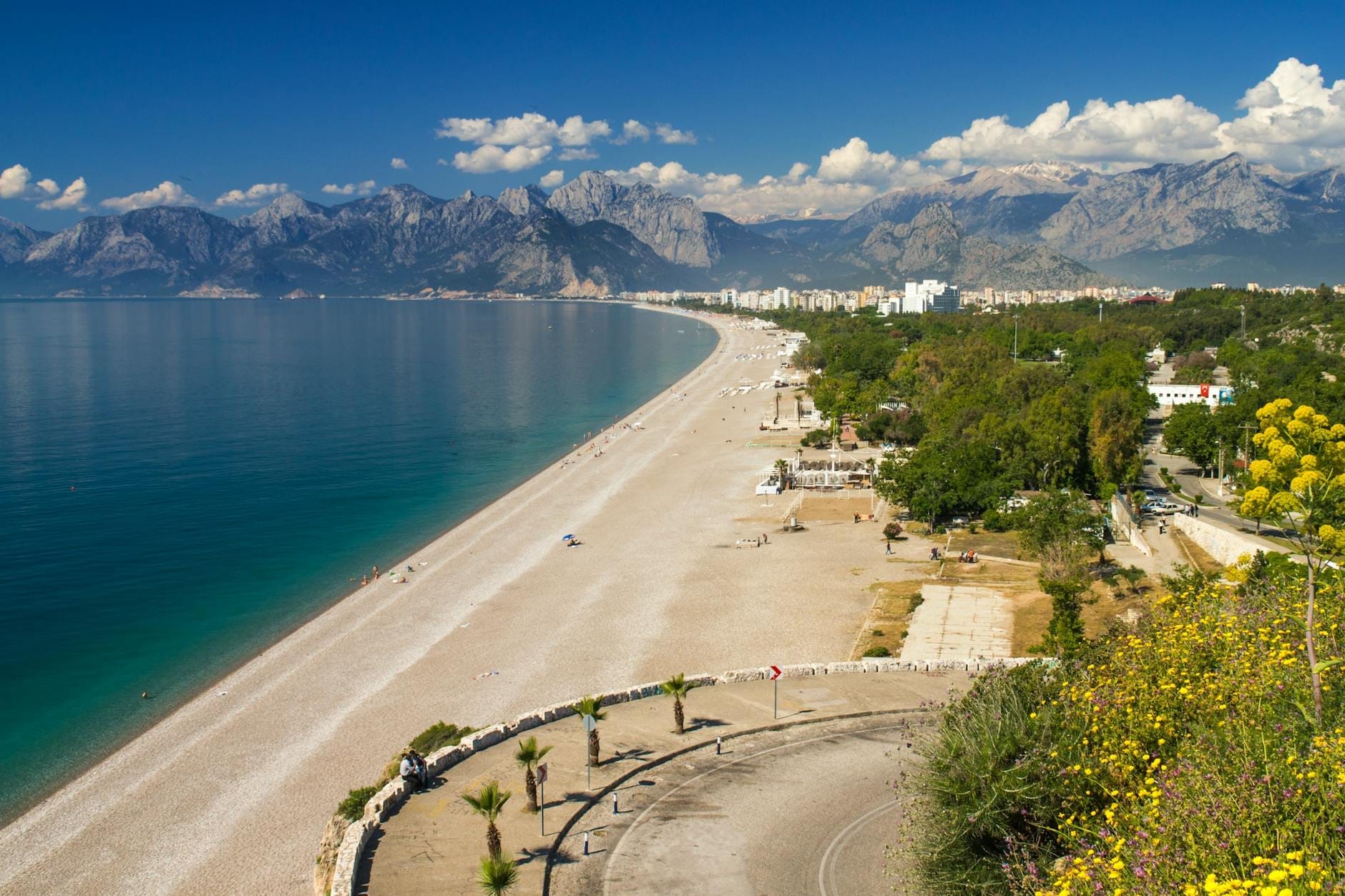 Una lunga distesa di sabbia della spiaggia di Lara ad Antalya, con acque azzurre e limpide, aree verdi e montagne imponenti sotto un cielo luminoso, vista dall'alto.